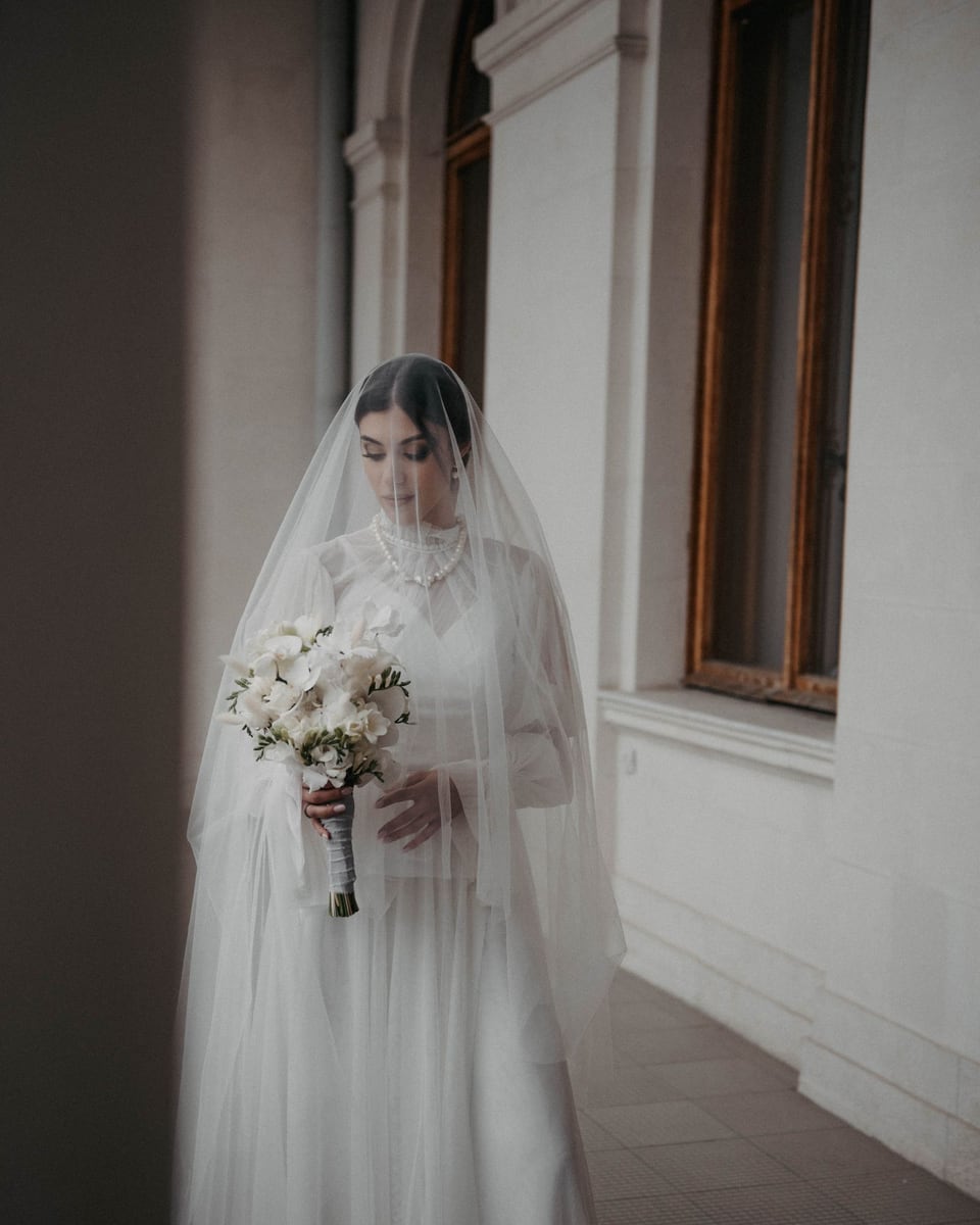 Illustrative photograph of a bride in a white gown and veil holding a bouquet in an airy interior.